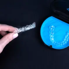 Dłoń trzyma przezroczysty aparat Invisalign. Obok, w niebieskim etui, leży kolejny.