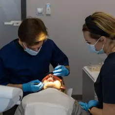Dentysta pracuje przy otwartych ustach pacjentki, która zastanawia się, ile trzyma znieczulenie u dentysty. Asystentka obserwuje zabieg.