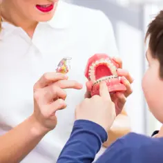 Ortodontka pokazuje chłopcu model szczęki z aparatem, wyjaśniając, jak skorygować krzywy zgryz.