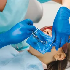 Dentysta zakłada koferdam, izolując pole zabiegu. Pacjentka leży na fotelu, ma założone okulary ochronne.