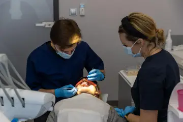 Dentysta pracuje przy otwartych ustach pacjentki, która zastanawia się, ile trzyma znieczulenie u dentysty. Asystentka obserwuje zabieg.