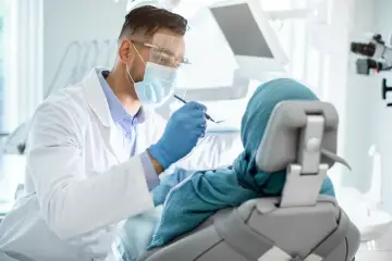 Lekarz stomatolog w masce i fartuchu bada pacjentkę w fotelu. Nawet przy łagodnym podejściu, dentofobia może być trudna do pokonania.
